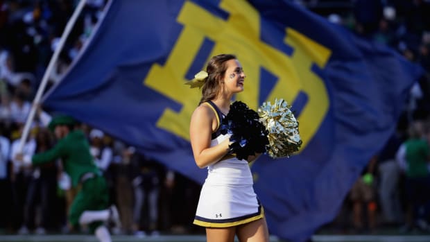 A Notre Dame cheerleader holding pompoms with the Notre Dame flag in the background.