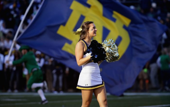 A Notre Dame cheerleader holding pompoms with the Notre Dame flag in the background.
