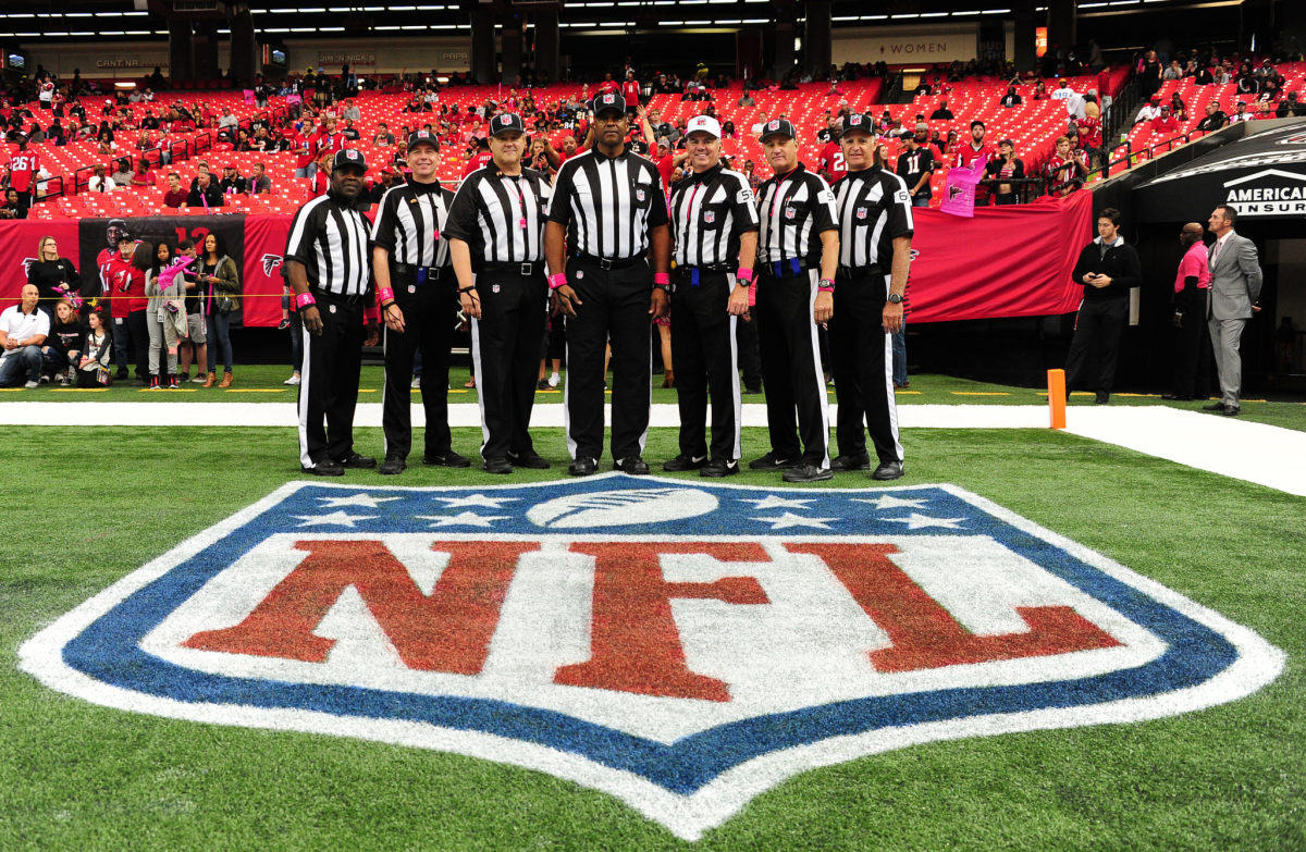 Bill Vinovich and other referees stand on field around NFL logo before a game.
