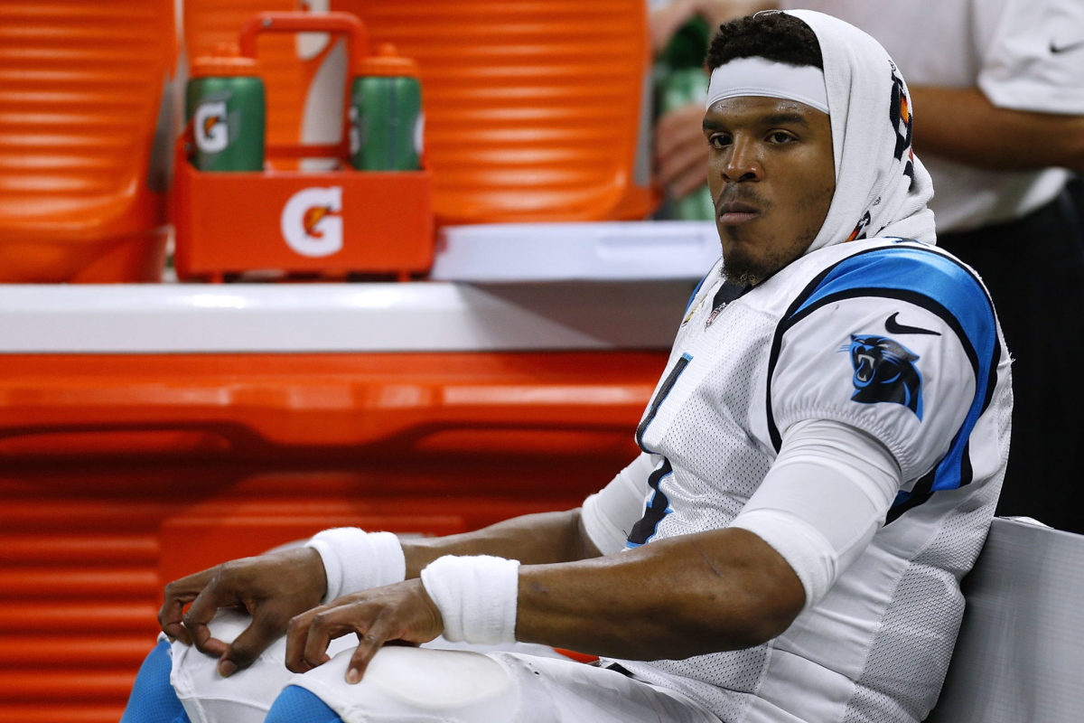 Cam Newton wearing a headband with a towel on his head on the sidelines .
