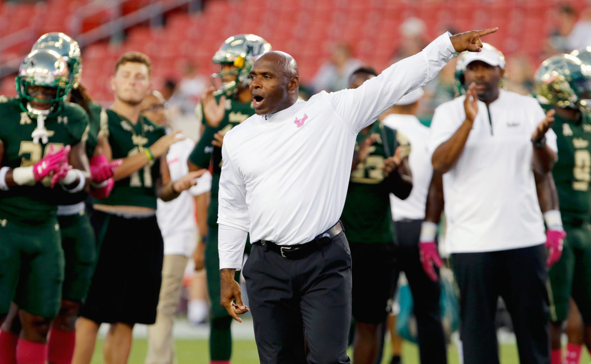 Charlie Strong pointing to the left while wearing a white turtleneck with a pink USF logo on it.