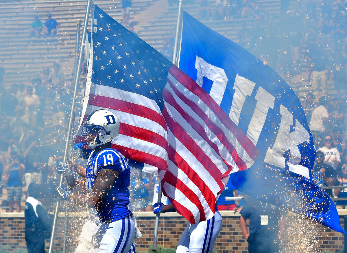 Duke football players takes the field carrying American Flag.